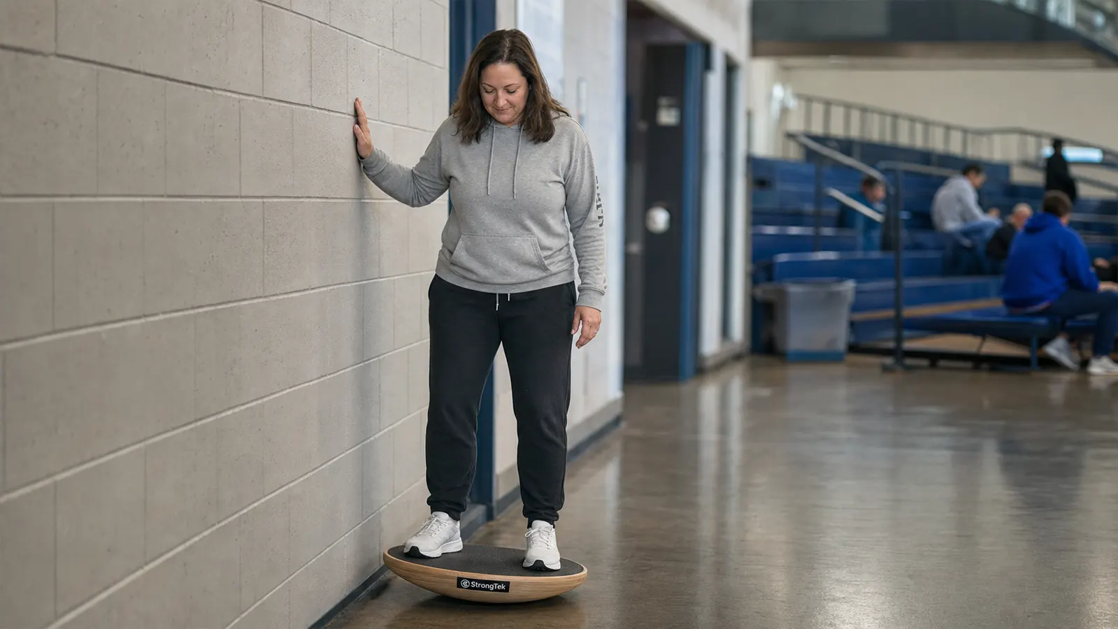 A teenage wrestler standing on a wooden rocker balance board in a gym hallway, one hand resting near a wall for balance