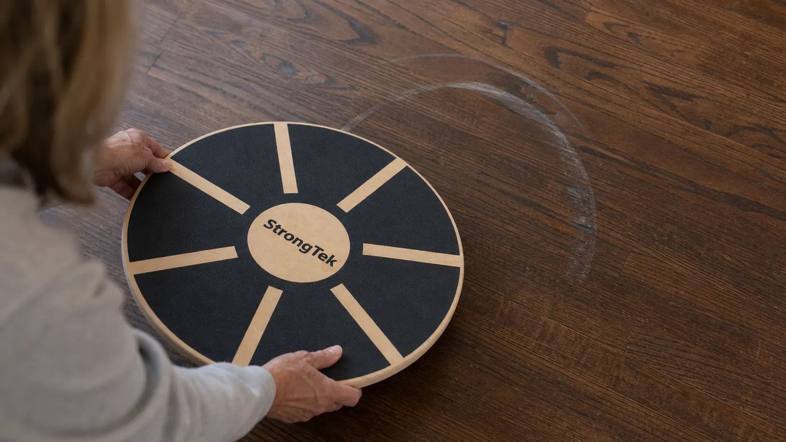Close-up of a hand pointing to a faint scuff mark on a hardwood floor next to the wooden balance board