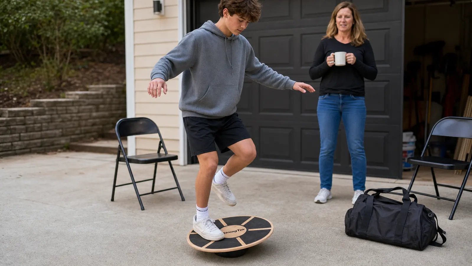 A teenage wrestler standing on one leg on a round wooden wobble balance board in a driveway, arms out for balance