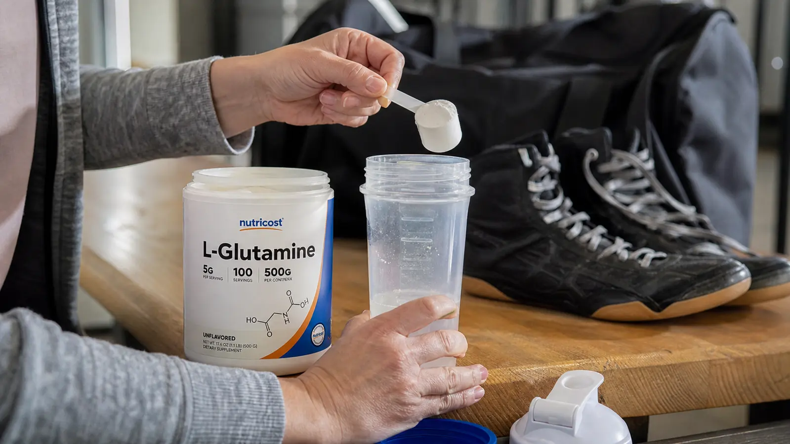 A hand scooping unflavored powder from the Nutricost L-Glutamine tub into a shaker bottle after practice