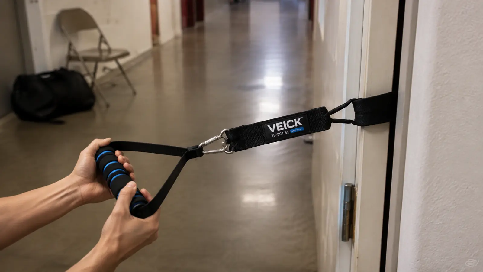 Hand gripping the molded handle of a VEICK resistance band with the door anchor wedged into a rec center hallway doorframe